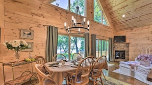 A wooden cabin interior with a dining table set for four, surrounded by wicker chairs, a chandelier, and a fireplace. - Spacious Pearl Lake Retreat w/ Yard & Private Dock (Kimball)