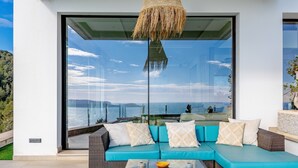 Un área de estar moderna al aire libre con un amplio ventanal que da a una vista panorámica del mar y las colinas.