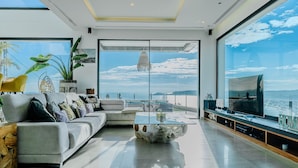 Una sala de estar moderna con un sofá grande, una mesa de centro y un televisor. Una ventana amplia ofrece vistas al mar y al paisaje urbano.