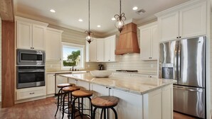 Uma cozinha moderna com armários brancos, eletrodomésticos de aço inoxidável, uma ilha grande com banquetas e iluminação pendente.