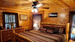 A wooden cabin room with a bed, a desk, a chair, a ceiling fan, and a window with curtains. - The Wild Buck, A Secluded Cabin that Borders the National Forest (Broken Bow)