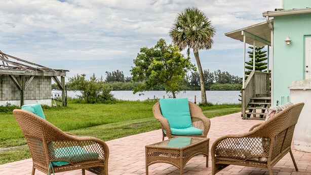 Wicker furniture with cushions, a glass table, and a view of a lake and greenery. - Riverview cottage (Grant-Valkaria)