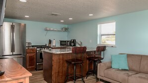 A compact kitchen with a central island, bar stools, a sofa, and a window with blinds. - Riverview cottage (Grant-Valkaria)