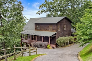 Een houten chalet met twee verdiepingen en een veranda, omringd door bomen en een verharde oprit.