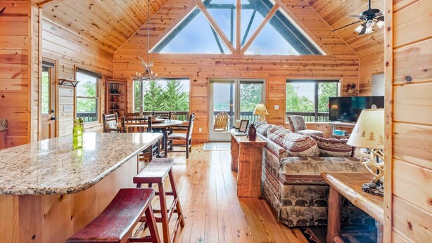 Una cabaña espaciosa con techos altos, un amplio comedor, una sala de estar acogedora con sofá y televisor, y grandes ventanas que ofrecen vistas al exterior.