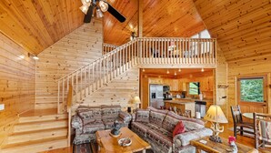 Interior de una cabaña de madera con una zona de estar central, una cocina y una escalera que sube a un altillo.