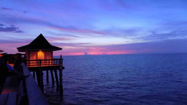 Un muelle de madera que lleva a una pequeña estructura iluminada con techo de paja, con vista al mar tranquilo al anochecer.