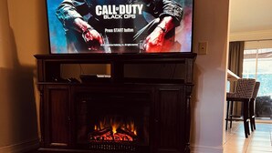 Una sala de estar con un televisor de pantalla plana mostrando 'Call of Duty Black Ops', una chimenea y controles de videojuegos sobre una mesa de vidrio.