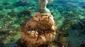 A statue of a woman on a rock by a clear blue lake with buildings in the background.