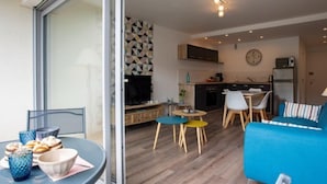A modern living room with a blue sofa, a dining table with chairs, a kitchen area with cabinets and appliances, and a large sliding glass door leading to an outdoor area. - Les Gemeaux by Interhome (Quiberon)