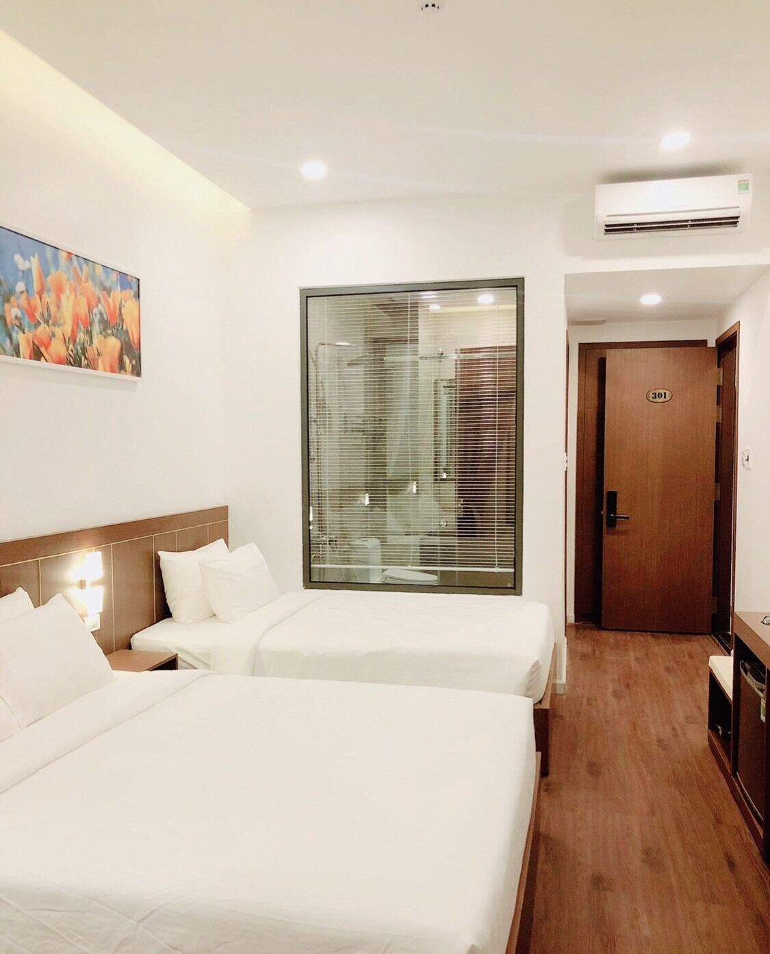Habitación de hotel con dos camas, un ventanal con persianas, un equipo de aire acondicionado y una puerta de madera con el número 301.