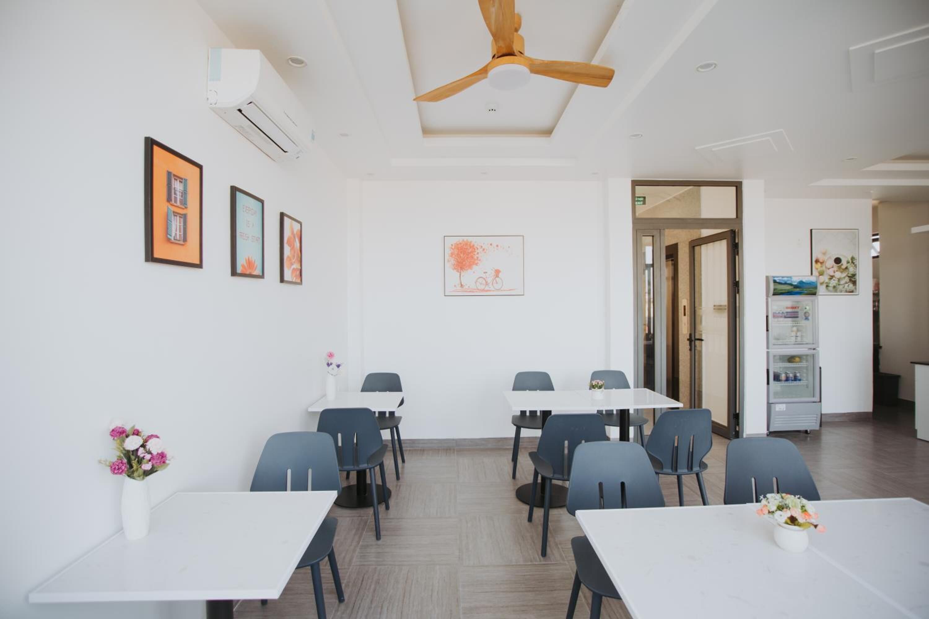 Un comedor moderno con mesas y sillas blancas, obras de arte enmarcadas en las paredes y un ventilador de techo.