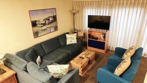 Een woonkamer met een bank, een blauwe fauteuil, een salontafel, een televisie en een schilderij aan de muur.
