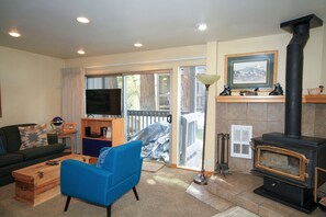 A living room with a sofa, a blue armchair, a wooden chest, a television, a fireplace, and a sliding glass door leading to an outdoor area. - Sierra Park Villas #20 (Mammoth Lakes)