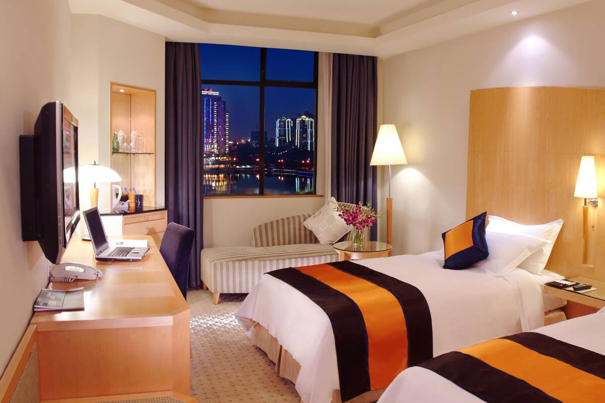 A hotel room with two beds, a desk, a television, and a view of a cityscape at night.