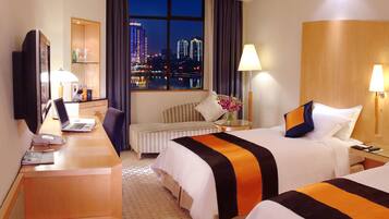 A hotel room with two beds, a desk, a television, and a view of a cityscape at night.