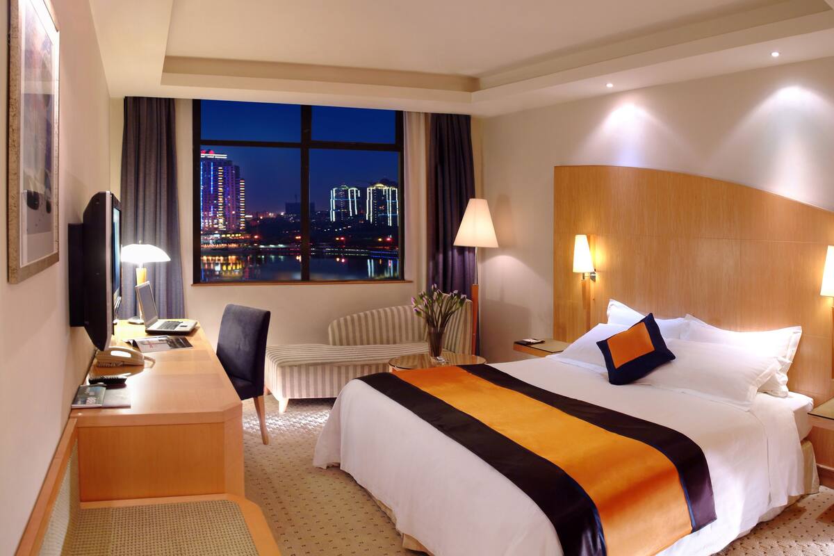 A hotel room with a bed, desk, chair, and a view of a cityscape at night.