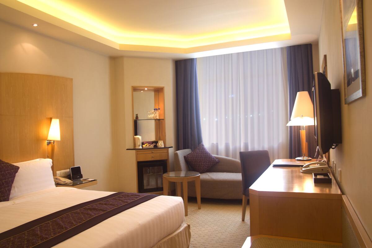 A hotel room with a large bed, a desk with a telephone, a chair, a small table, a lamp, a mirror, and a TV on the wall.