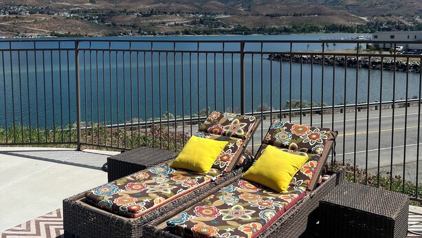 A balcony with wicker furniture and yellow cushions, overlooking a lake and hills. - Floating Over The Water! (Chelan)
