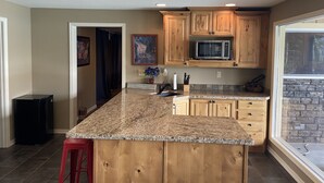 A kitchen with wooden cabinets, a granite countertop, a microwave, and a window with a stone wall. - Floating Over The Water! (Chelan)