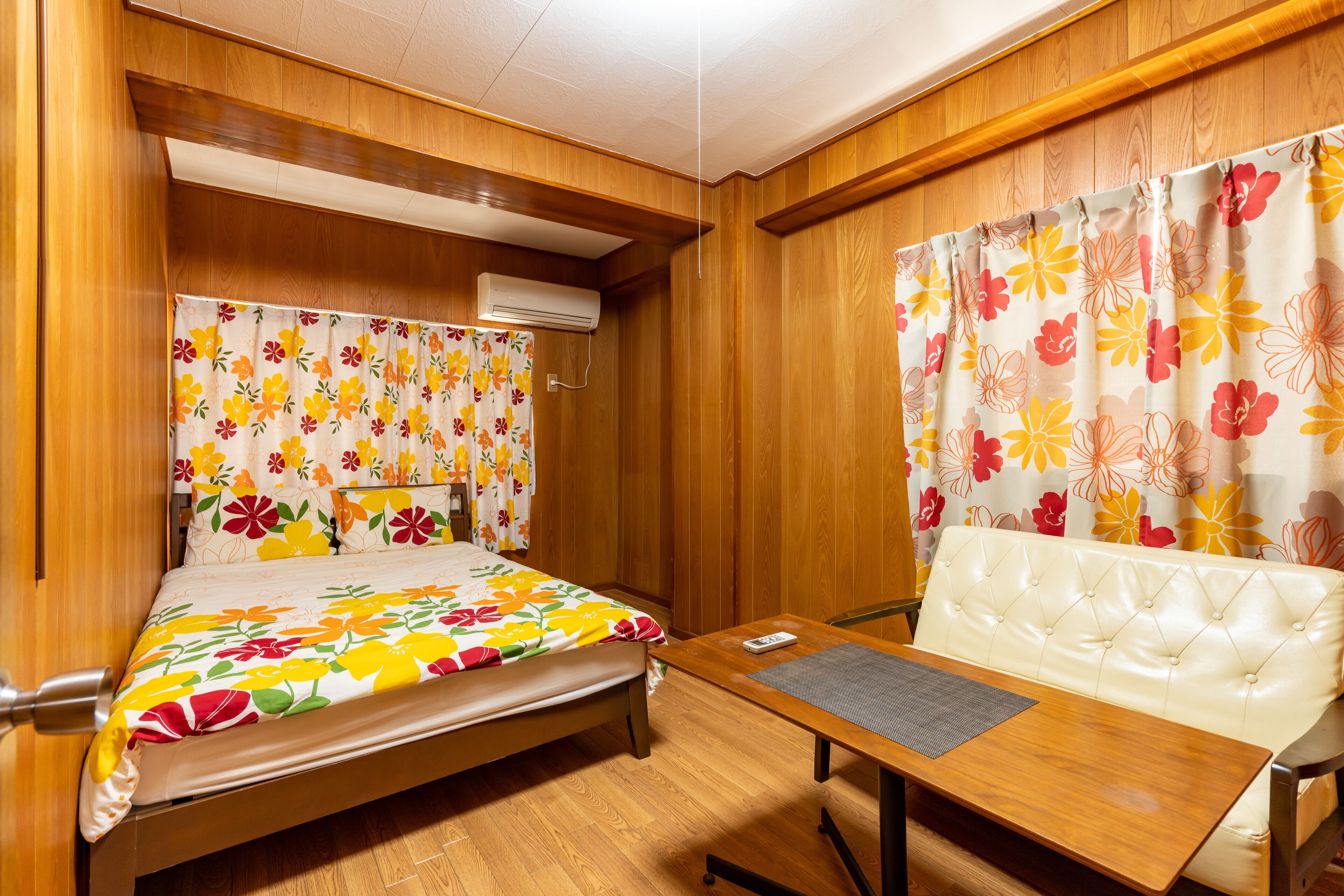Ett rum med träpanel på väggarna, en säng med ett blommigt överkast, en vit stoppad soffa och ett litet bord med en matta.