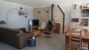 Uma sala de estar com sofá marrom, TV, parede de pedra, lareira e área de jantar com cadeiras de madeira e uma mesa.