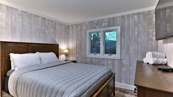 Une chambre à coucher avec une tête de lit en bois, un grand lit recouvert d’une couette grise, une table de chevet avec une lampe, une commode en bois, une fenêtre donnant sur des arbres et un téléviseur fixé au mur.