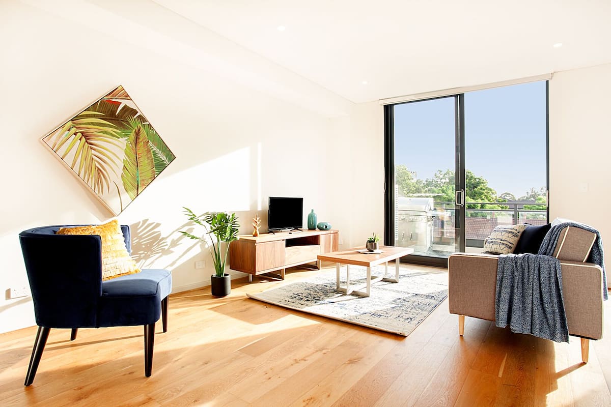 Ein modernes Wohnzimmer mit großem Fenster, einem Flachbildfernseher, einer Couch, einem Sessel und einem Couchtisch.