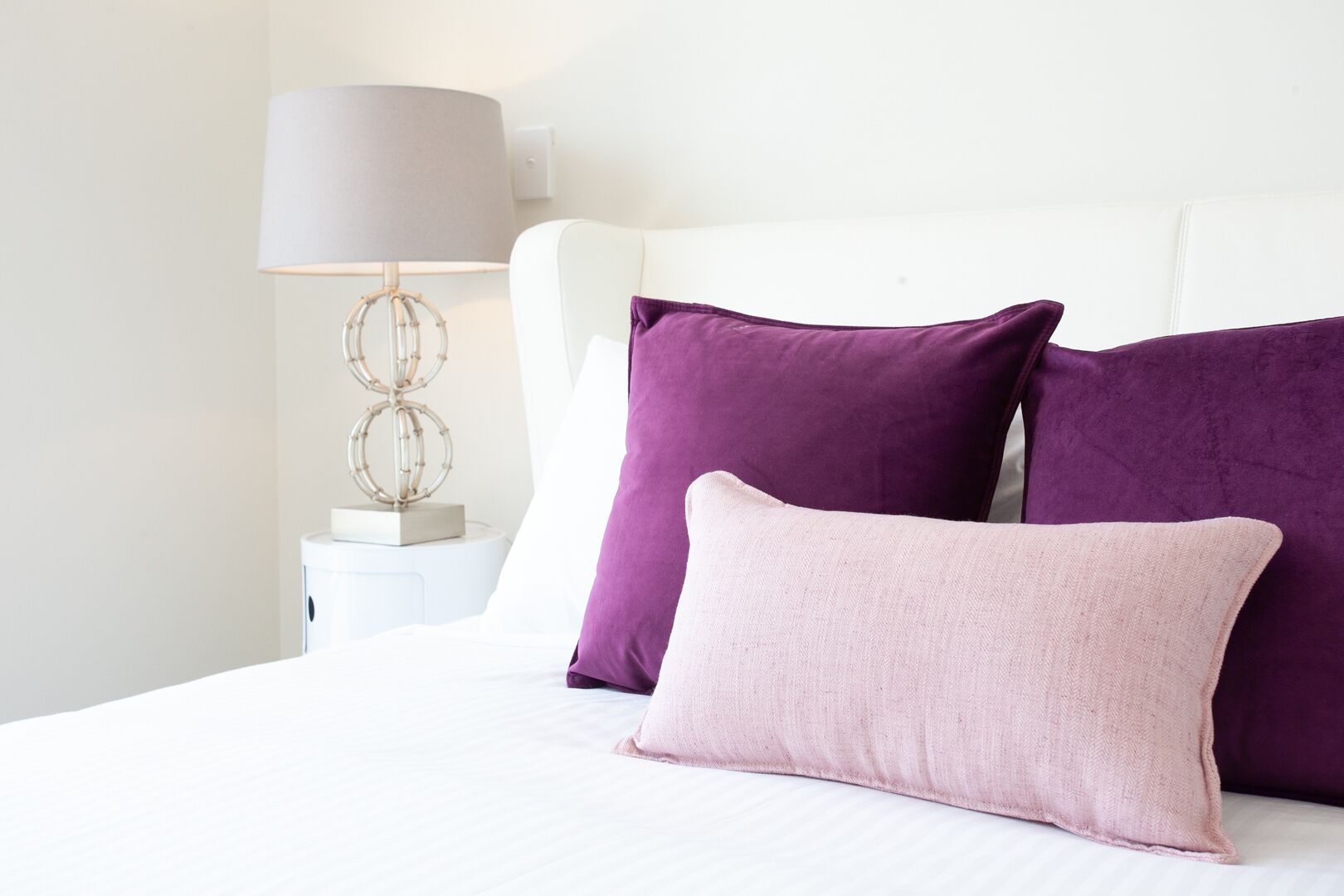 A neatly made bed with white bedding and three pillows in shades of purple and pink.