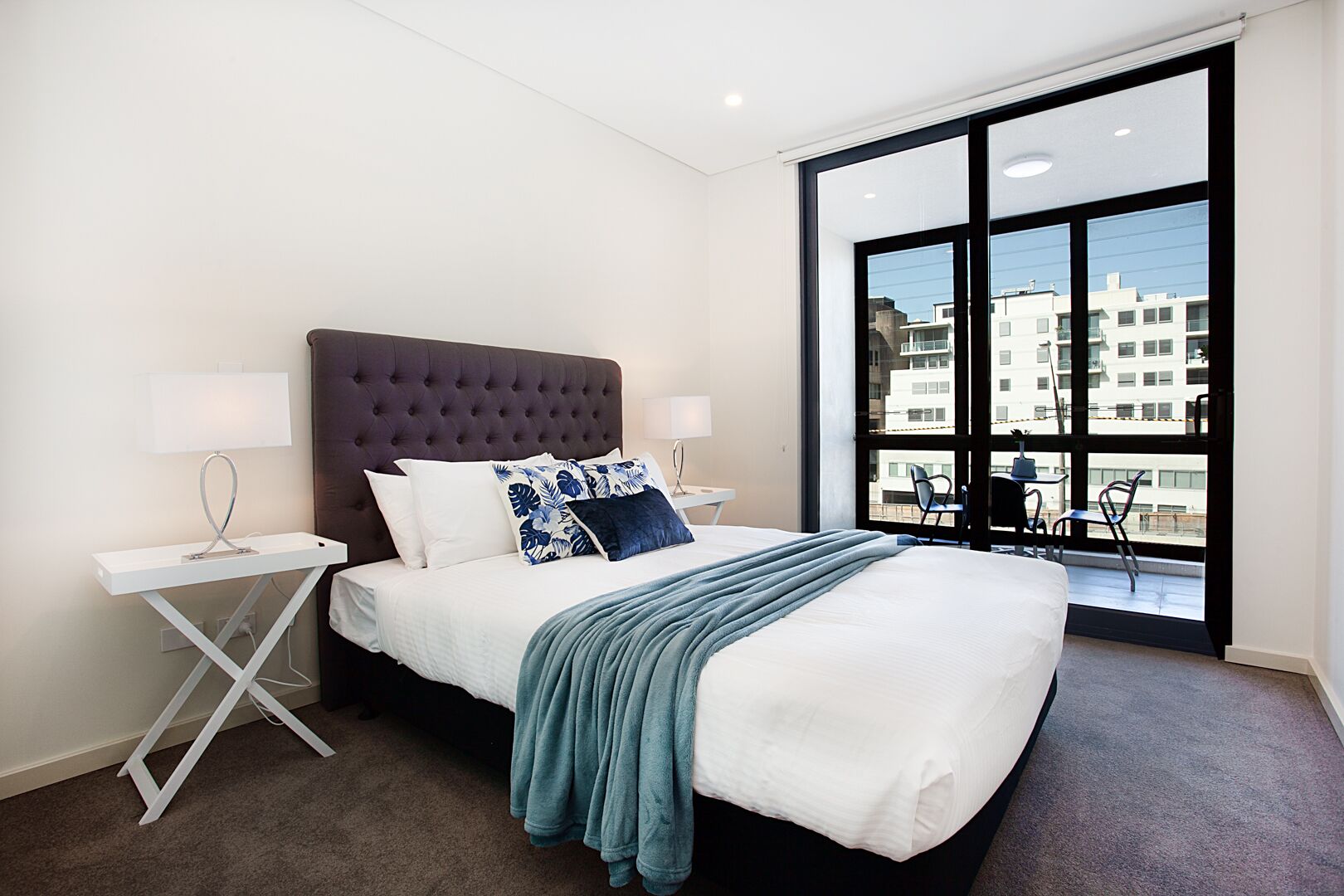 A modern hotel room with a large bed, two bedside tables, a window with a view of buildings, and a balcony with chairs.