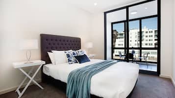 A modern hotel room with a large bed, two bedside tables, a window with a view of buildings, and a balcony with chairs.