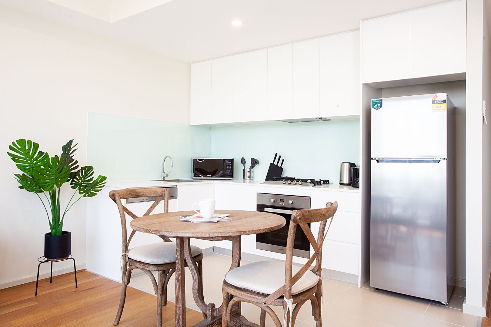 A modern kitchen with white cabinets, a stainless steel refrigerator, a microwave, and a stove. There is a dining table with chairs and a potted plant.