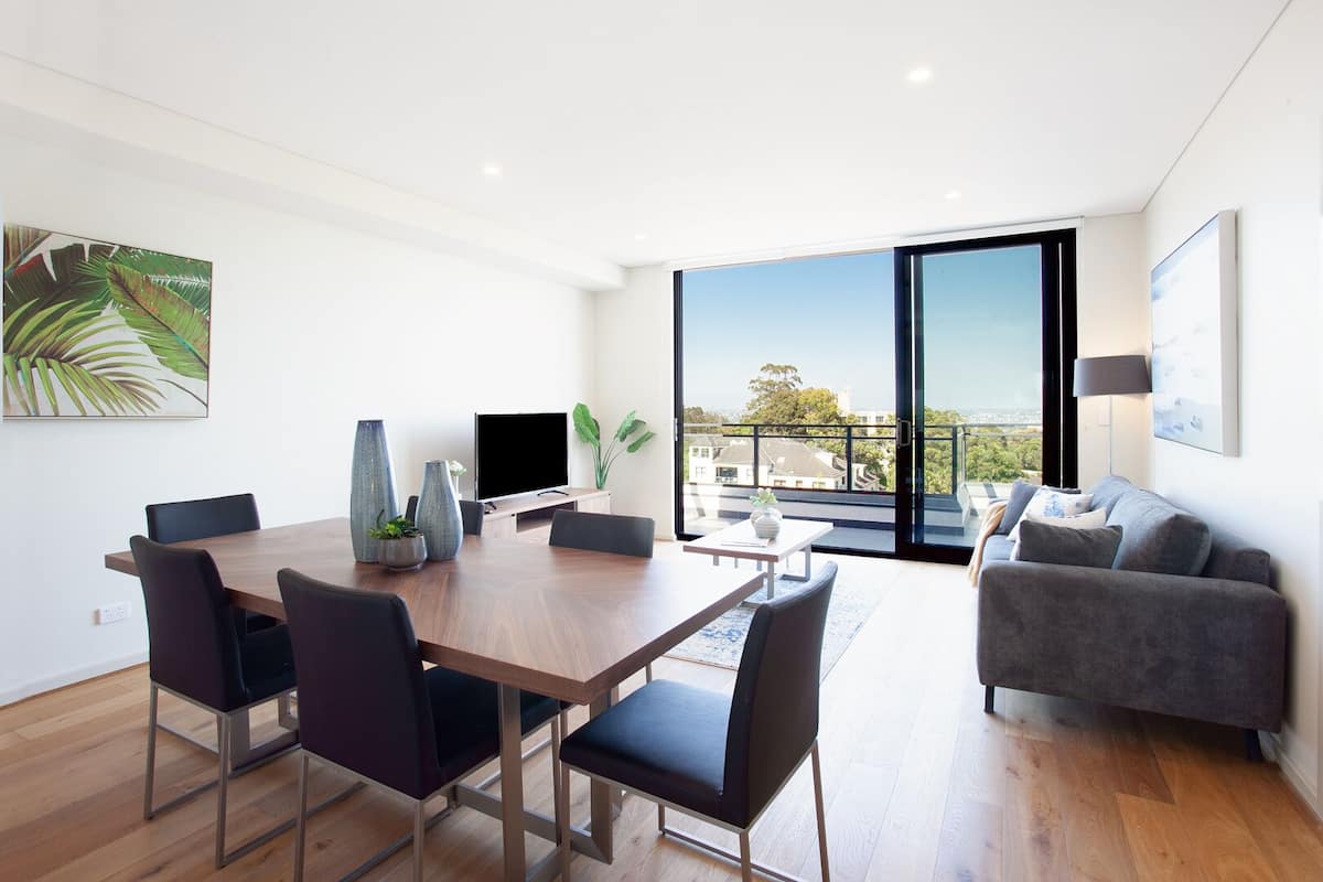 Ein modernes Wohnzimmer mit Essbereich, großen Fenstern und einem Balkon mit Blick auf die Stadt.