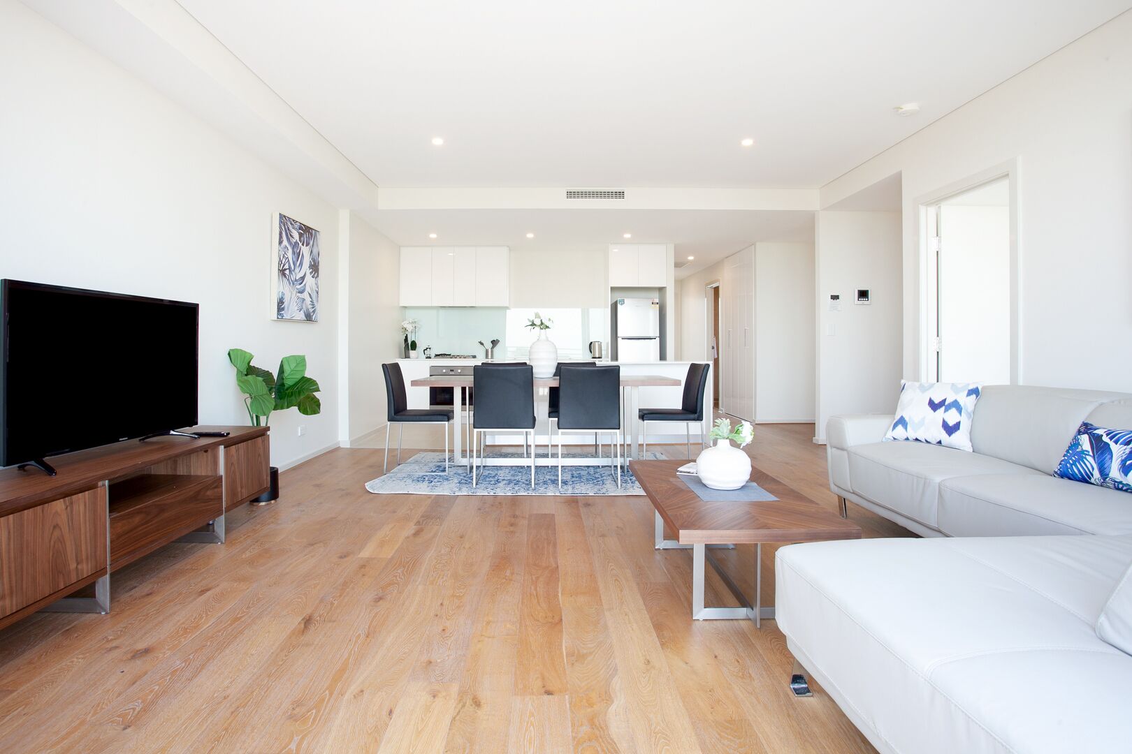 A modern living room with a flat-screen TV, wooden flooring, a white sofa, and a dining area with a kitchen in the background.