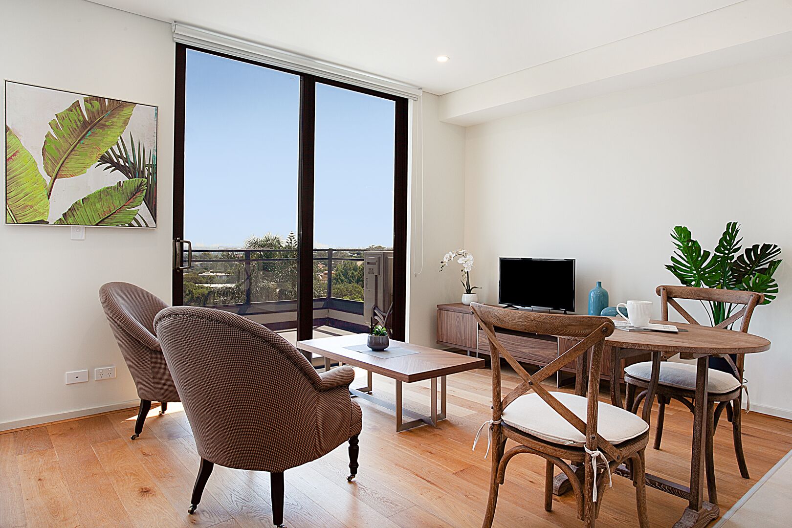 A spacious living room with a large window, a flat-screen TV, a dining table with chairs, and a potted plant.