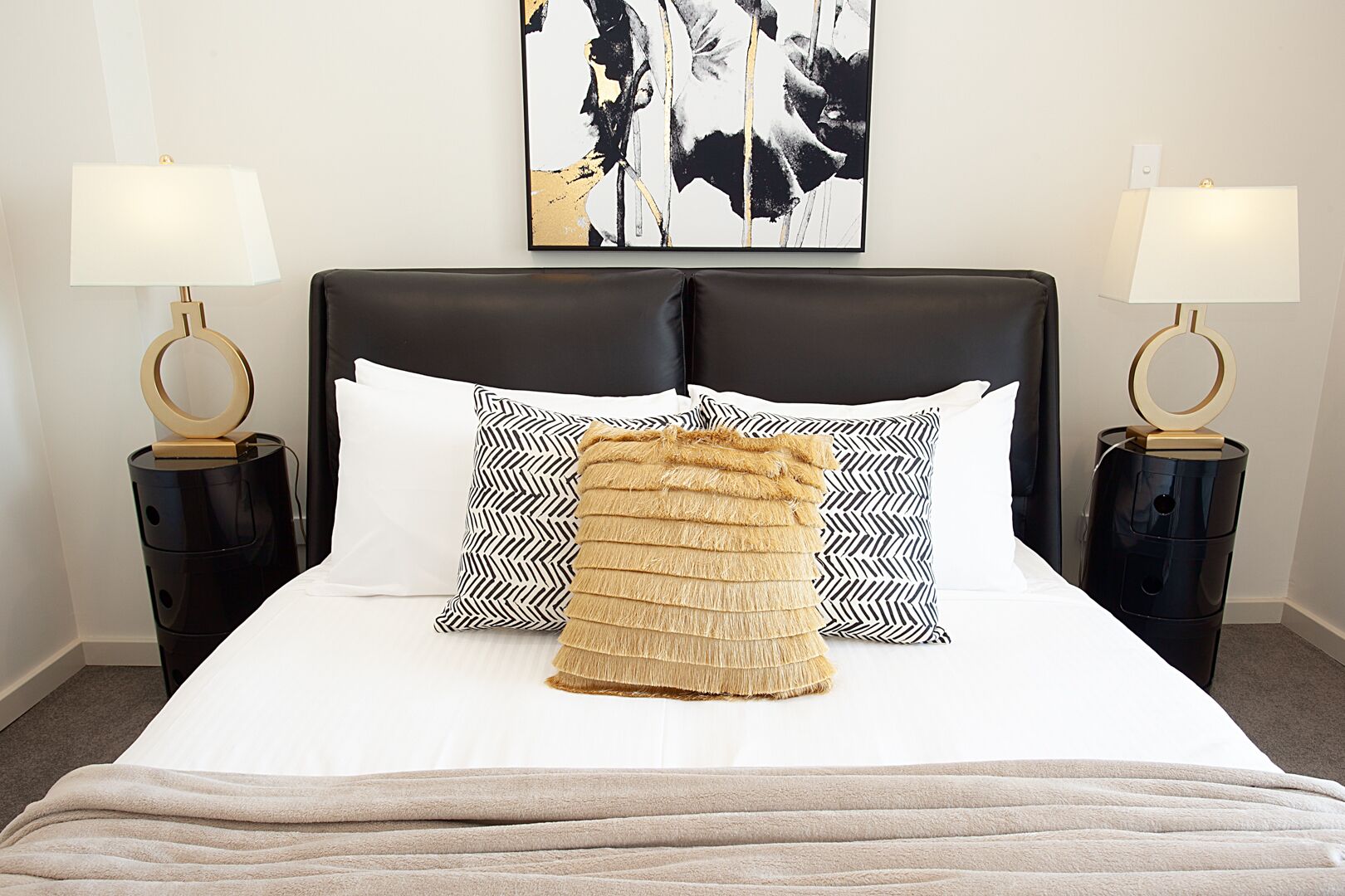 Un lit bien fait, avec une couette blanche, deux coussins à motifs noir et blanc, un plaid beige et une tête de lit noire. On y trouve deux tables de chevet avec des lampes, et une œuvre d’art murale au-dessus du lit.