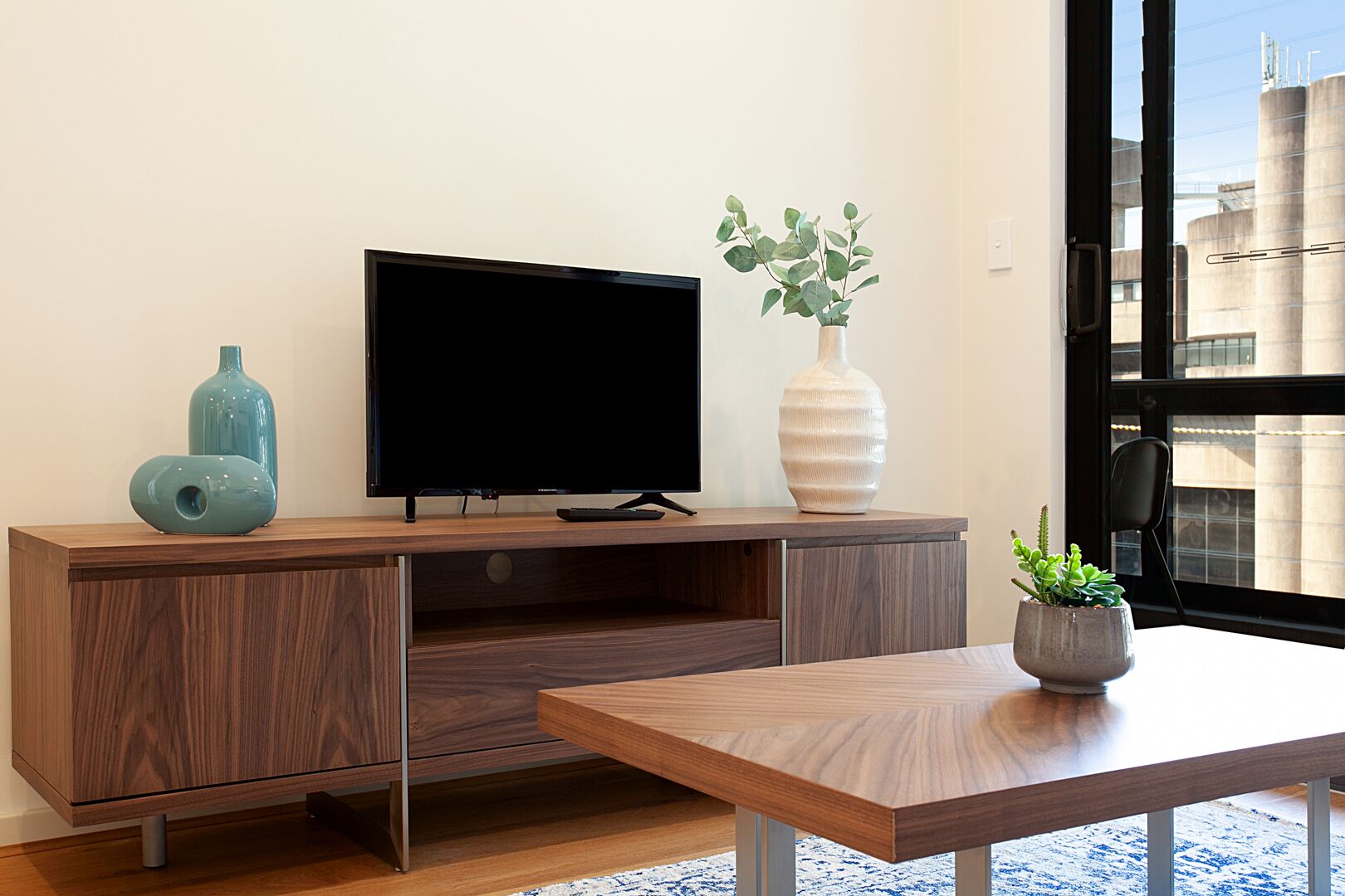 Una sala de estar moderna con un mueble bajo para televisor de madera, un televisor de pantalla plana, un florero con plantas verdes y una pequeña mesa con una planta en maceta.