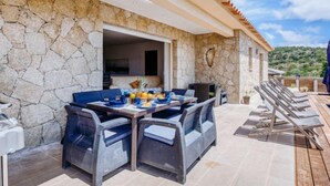 An outdoor patio with a stone wall, wicker furniture, and a view of the hills. - Villa Belios by Interhome (Porto Vecchio)