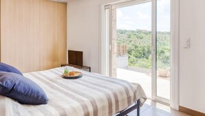 A bedroom with a bed, a nightstand, a TV, and a large sliding glass door leading to a balcony with a view of greenery. - Villa Belios by Interhome (Porto Vecchio)