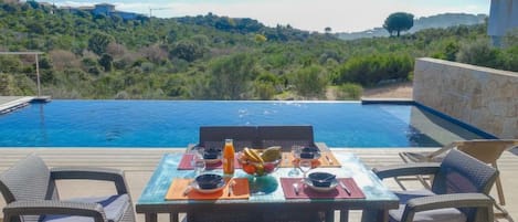 An outdoor dining area with a table set for two, overlooking a swimming pool and a scenic hillside view.