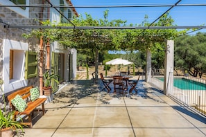Une terrasse avec une table et des chaises, un banc, une pergola couverte de plantes grimpantes, une piscine et un mur en pierre.