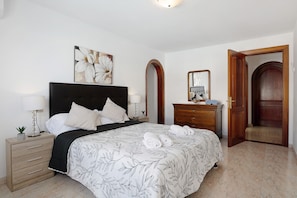 A bedroom with a bed, bedside table, dresser, mirror, and an open door leading to another room.