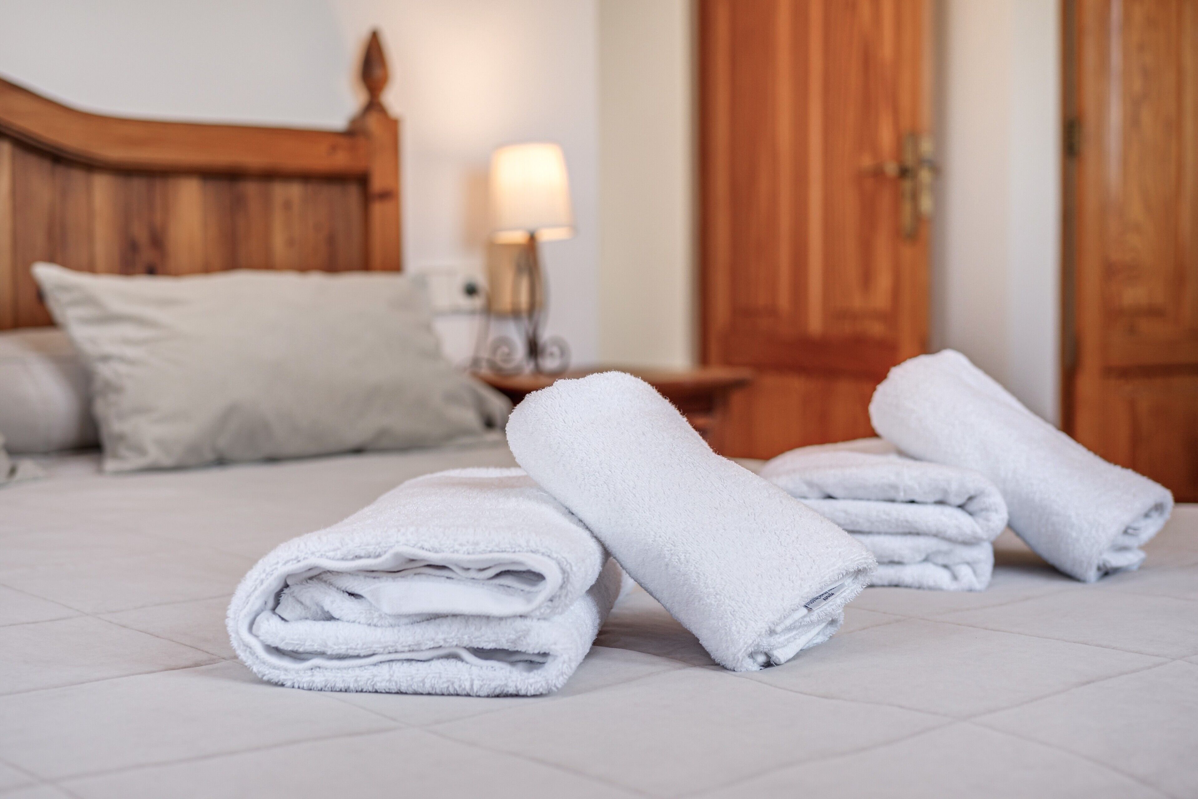 A neatly made bed with folded white towels, a wooden headboard, a bedside lamp, and a wooden door in the background.