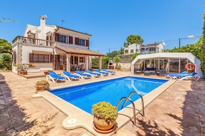 Une piscine avec des chaises longues, un espace de détente couvert et un bâtiment doté d’un balcon.