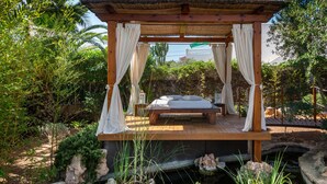 A wooden four-poster bed with white curtains, set on a wooden deck with a stone wall and greenery in the background.