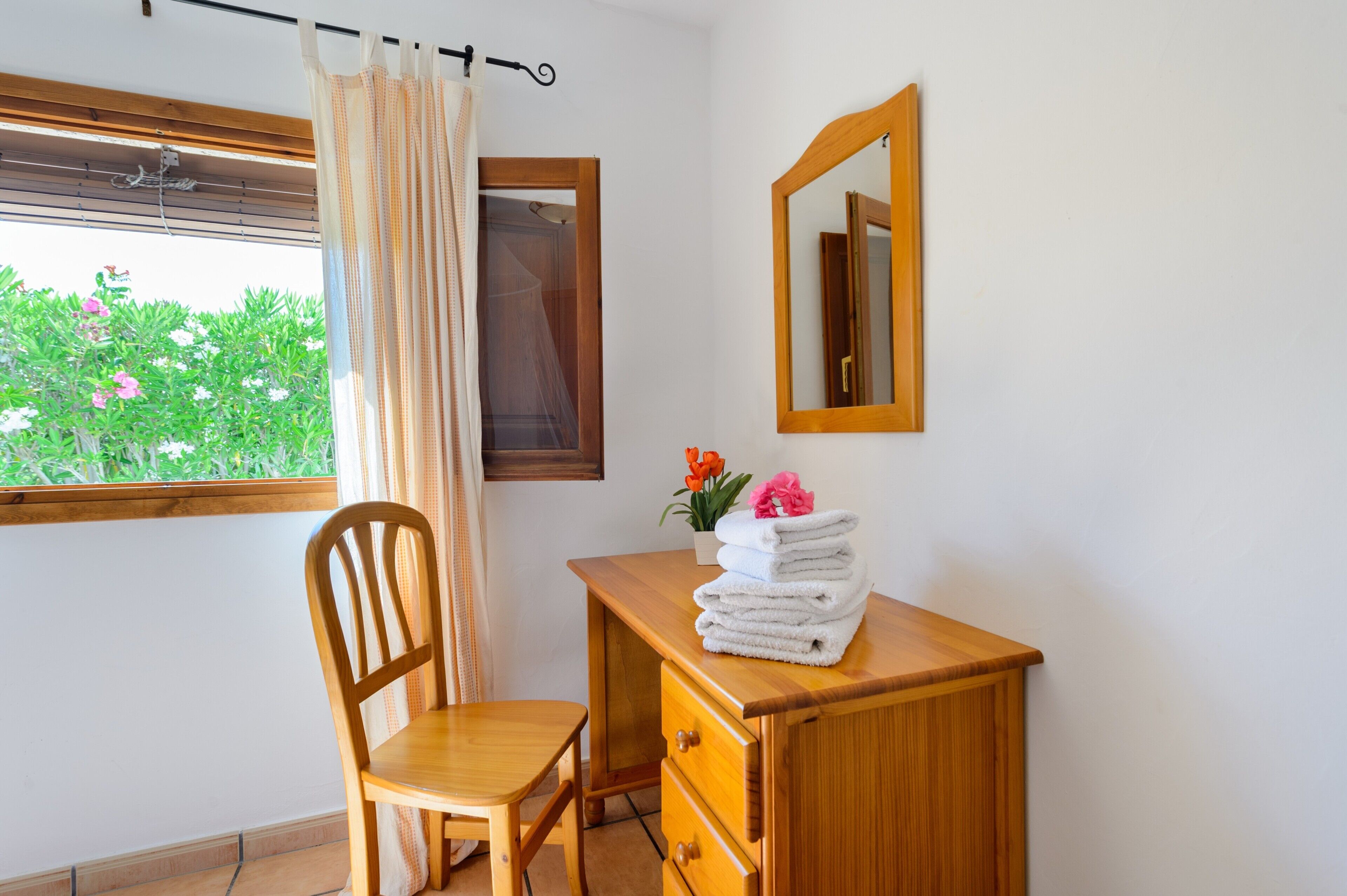 Een kamer met een houten bureau, een stoel, een raam met uitzicht op groen, een spiegel en een gordijn.