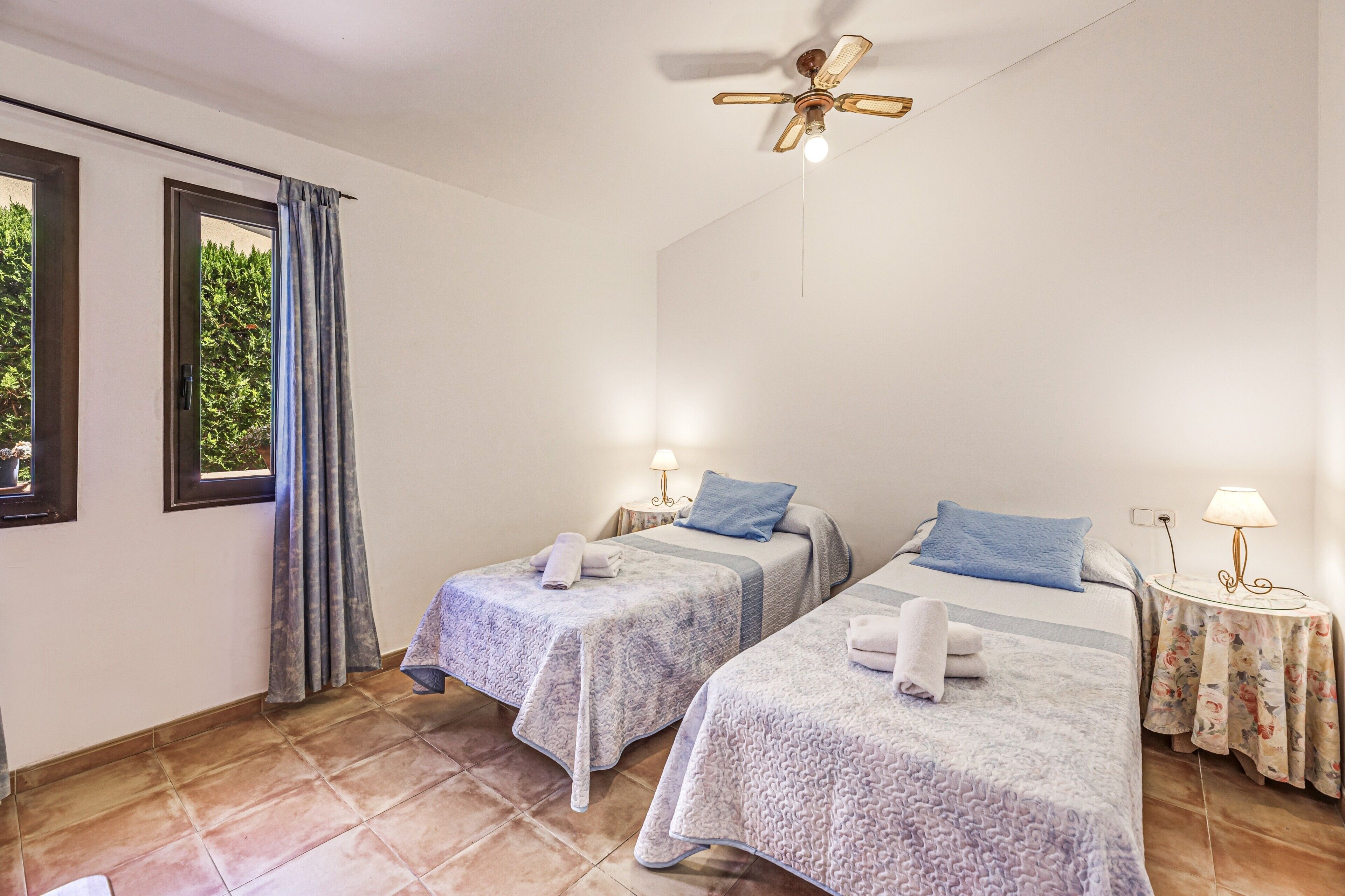 A room with two beds, a ceiling fan, a window with curtains, and a nightstand with a lamp.