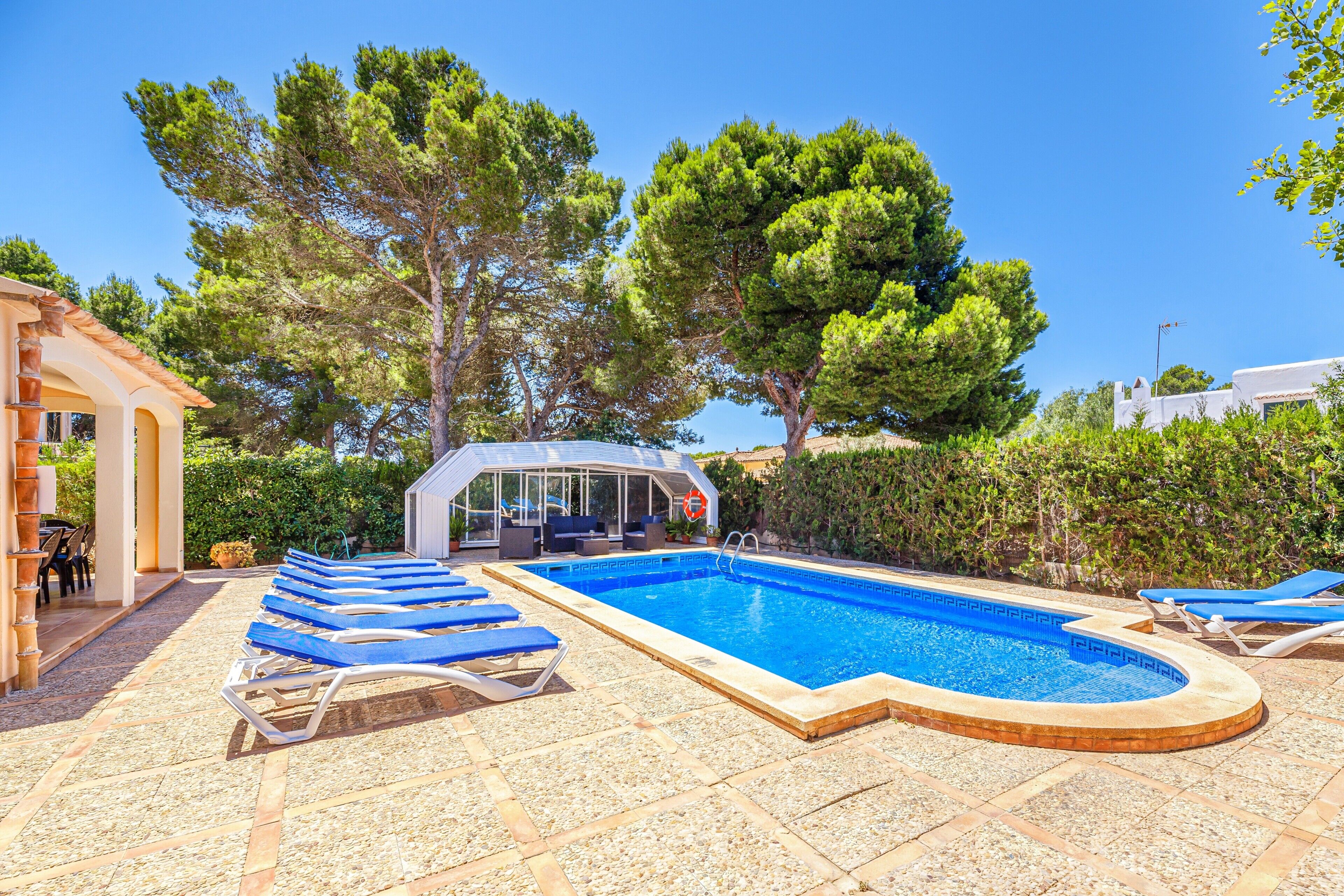 Ein Swimmingpool mit Liegestühlen, einer überdachten Sitzgelegenheit und Bäumen im Hintergrund.