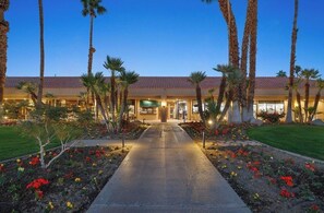 A hotel entrance with a pathway leading to the building, surrounded by palm trees and flower beds.