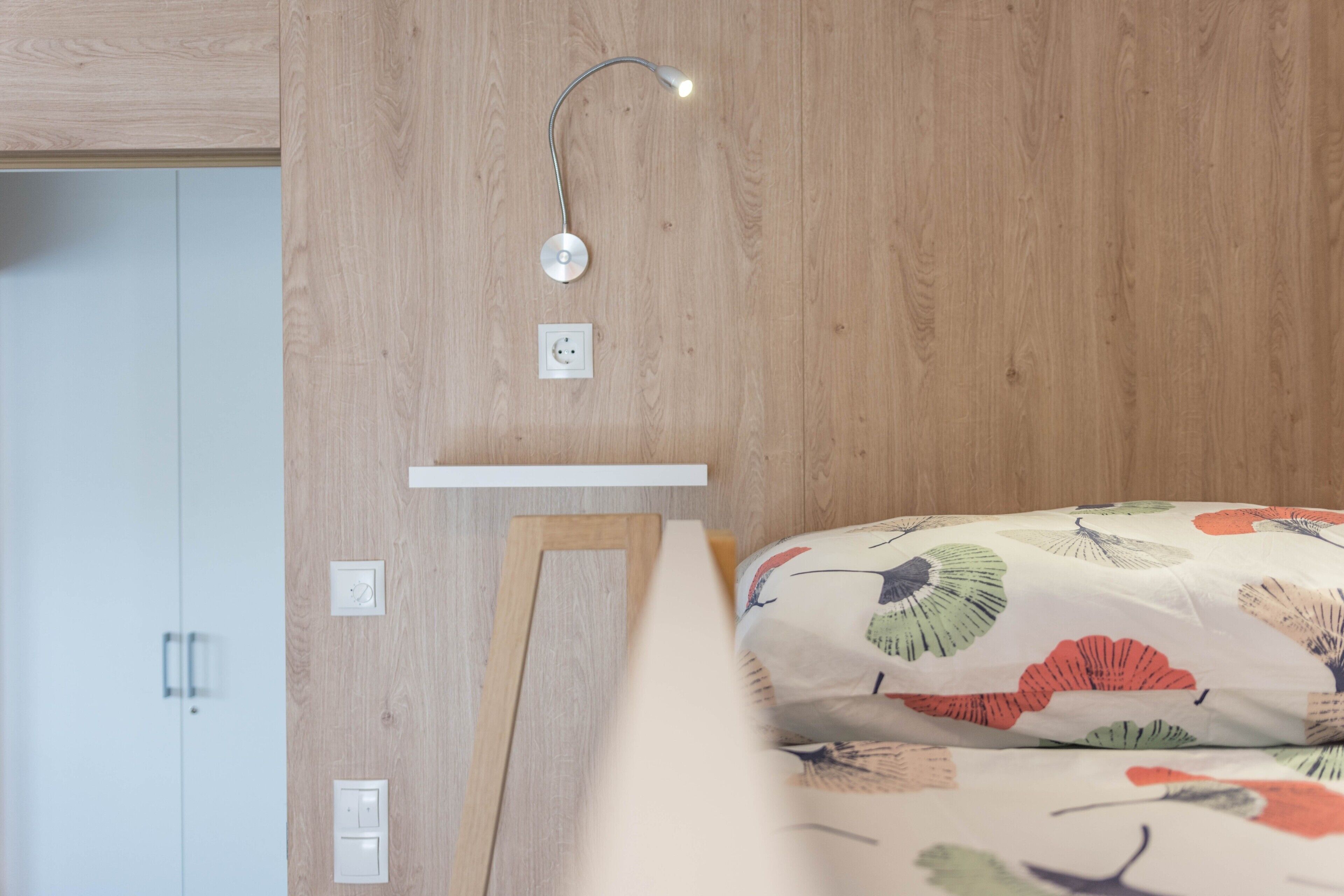 Une chambre avec un mur en bois, une table de chevet, une lampe et un lit recouvert d’une couvre-lit à motifs floraux.